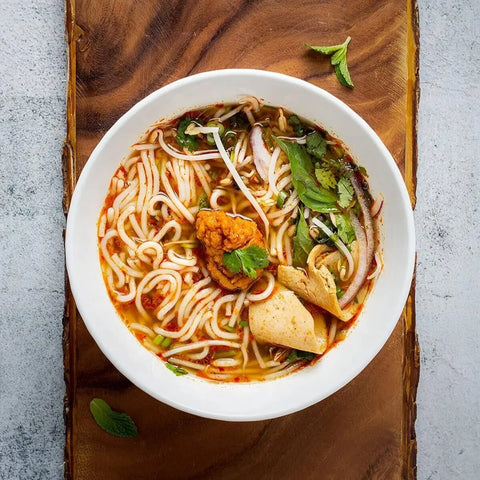 A white bowl of noodle soup with herbs, fish cakes, veggies, and chili paste sits on wood. The reddish broth is enhanced by Gustus Vitae’s Fried Rice Seasoning Collection, while fresh greens garnish the vibrant dish.
