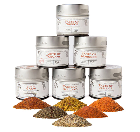 Six tins from the Gustus Vitae Cuisines of the World Gourmet Seasonings Collection are stacked in a pyramid, with vibrant piles of ground spices displayed in front on a white background.