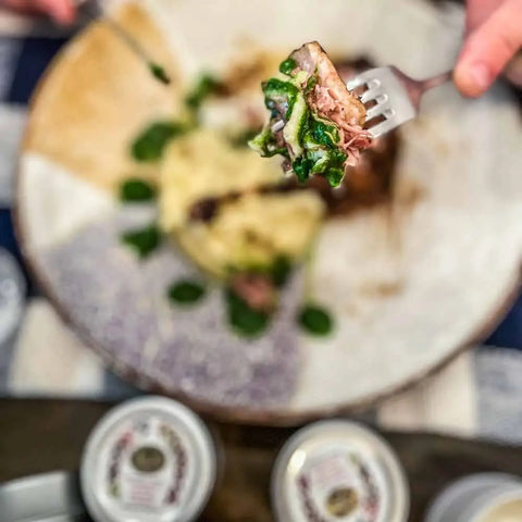 A fork holds a bite of greens and meat over mashed potatoes and sauce, with three tins from the Gustus Vitae Cuisines of the World Gourmet Seasonings Collection visible in the foreground.