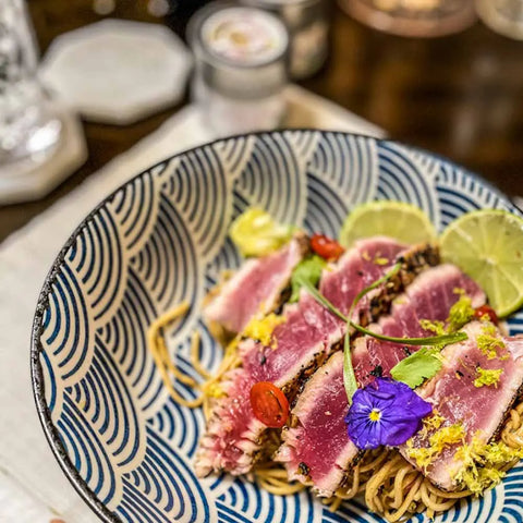 Seared tuna on noodles with lime, cherry tomatoes, greens, and a purple edible flower shines in a patterned bowl—each bite elevated by Gustus Vitae’s Cuisines of the World Gourmet Seasonings Collection. A glass and dining items blur softly in the background.