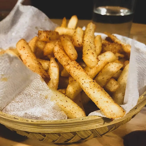 A basket of fries seasoned with Gustus Vitae’s Taste of Home – For Your Air Fryer (4 Pack) spice blend sits on a paper towel atop a wooden surface, with a glass of water in the background.