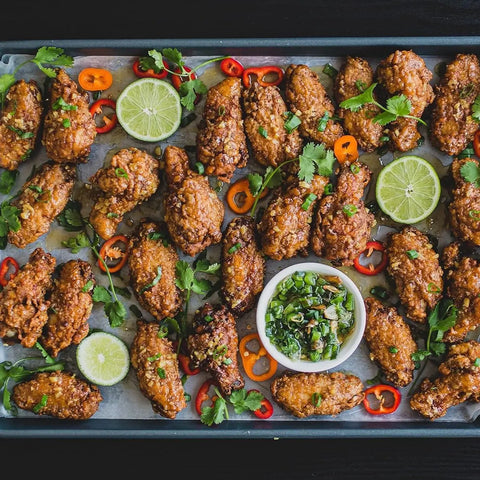 A tray of glazed chicken wings made with Gustus Vitae’s Taste of Home – For Your Air Fryer (4 Pack), garnished with cilantro, red and orange chili peppers, lime slices, and served with a green herb dipping sauce.