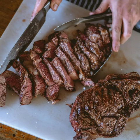A person expertly slices a juicy, grilled steak on a white cutting board, seasoned with Gustus Vitae’s Gourmet Backyard BBQ Favorites - 4 Pack, using a knife and tongs. Thick pieces reveal perfect grill marks and mouthwatering flavor.