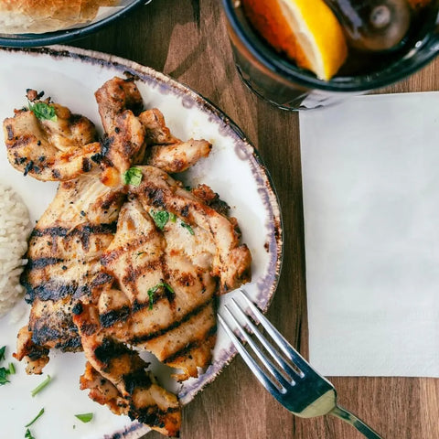 Enjoy a plate of grilled chicken seasoned with Gustus Vitaes Gourmet Backyard BBQ Favorites - 4 Pack, paired with white rice. A fork and a glass of iced drink with lemon rest beside it on a wooden table.