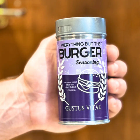 A hand holds a purple and silver “Gourmet Backyard BBQ Favorites - 4 Pack” by Gustus Vitae, featuring burger-themed accents—an artisan spice blend set that’s perfect for your BBQ seasoning collection.