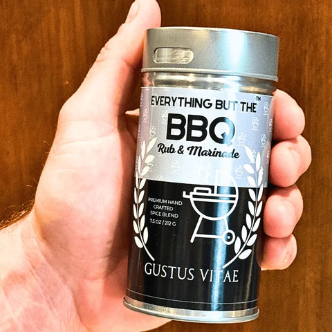 A hand holds a metal container from Gustus Vitaes Gourmet Backyard BBQ Favorites - 4 Pack, featuring a grill icon and white laurel leaves on a black label. The artisan spice blend container includes a shaker lid.