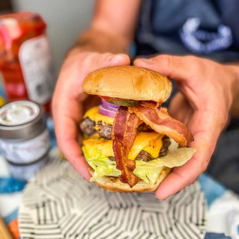 A person holds a cheeseburger loaded with bacon, lettuce, tomato, pickles, red onion, and cheddar in a bun. In the background are Gustus Vitae’s Everything but the Classic Cookout spice set of 4 and ketchup, suggesting handcrafted flavor.
