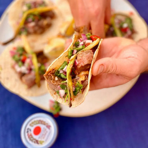 A hand holding a taco brimming with grilled meat and veggies hovers over a plate of more tacos, with Gustus Vitae’s Everything but the Fajita Night Collection | Set of 4 artisanal spice blends nearby on a blue tablecloth.