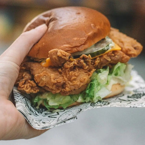 A hand holds a fried chicken sandwich on a soft bun with lettuce, pickles, and cheddar cheese, seasoned with Gustus Vitae’s Everything but the Food Truck Favorite Flavors Collection | Set of 4 for crave-worthy taste.