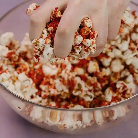 A hand grabs popcorn dusted with red seasoning from a glass bowl, featuring the Gustus Vitae Ultimate Popcorn Seasoning Set - Six Pack for an artisanal and elevated snacking experience.