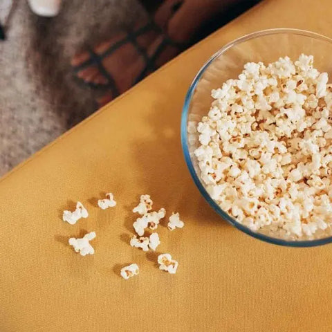 A glass bowl of popcorn, seasoned with Gustus Vitaes Ultimate Popcorn Seasoning Set - Six Pack, rests on a yellow surface, with some kernels spilled and sandal-clad feet visible in the carpeted background.