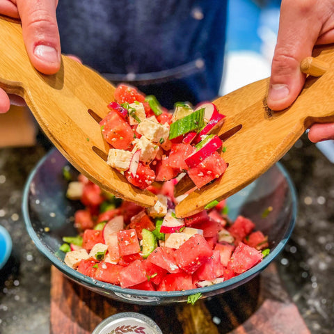 Close-up of hands tossing a watermelon salad with feta, cucumber, and red onion using wooden servers, enhanced with Gustus Vitaes Super Salad Seasonings Collection (Set of 4) for extra flavor.