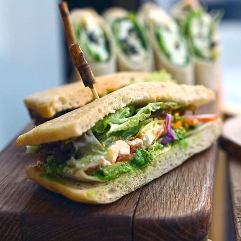 A fresh sandwich with lettuce, tomato, greens, and cheese on focaccia bread is secured with a wooden pick and enhanced by Gustus Vitae’s Super Salad Seasonings Collection | Set of 4, displayed on a wooden board. Rolled sandwiches blur in the background.