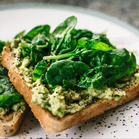 Two slices of whole wheat toast topped with mashed avocado, fresh spinach, and a dash of Gustus Vitae Super Salad Seasonings Collection, sprinkled with black pepper and served on a white plate.