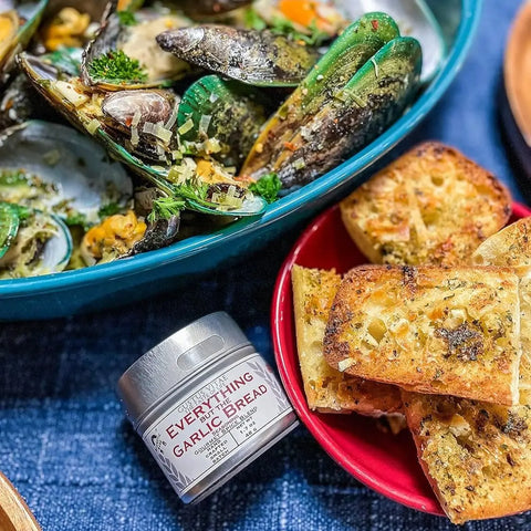 A bowl of seasoned mussels sits beside a red bowl of crispy garlic bread, with a jar of Gustus Vitae’s Ultimate Everything but The...Everything Seasonings Collection - Complete 12 Pack Set between them on a blue tablecloth.
