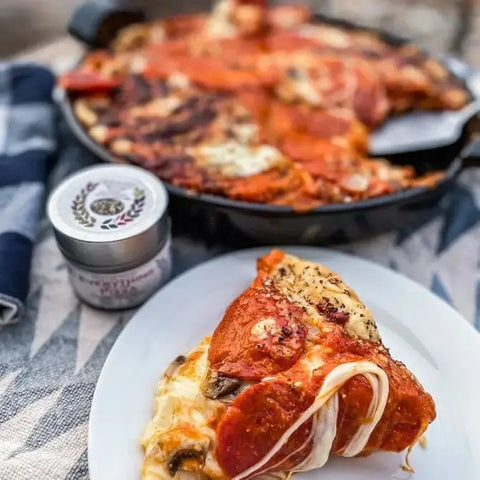 A cheesy slice of pepperoni pizza sits on a white plate before a cast iron skillet with more pizza and a jar from Gustus Vitaes Ultimate Everything but The...Everything Seasonings Collection 12 Pack Set, all atop a patterned tablecloth.