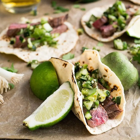 Three soft steak tacos with fresh green salsa and cilantro rest on parchment, enhanced by Gustus Vitae Luxury World Seasonings - Gourmet 12 Pack Collection. Zesty limes add brightness to this flavorful, colorful dish.