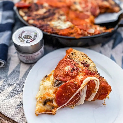 A cheesy pepperoni pizza slice stretches on a white plate beside a jar from Gustus Vitaes Everything but the Weekender Collection (Set of 4). More pizza can be seen in a cast iron skillet in the background.