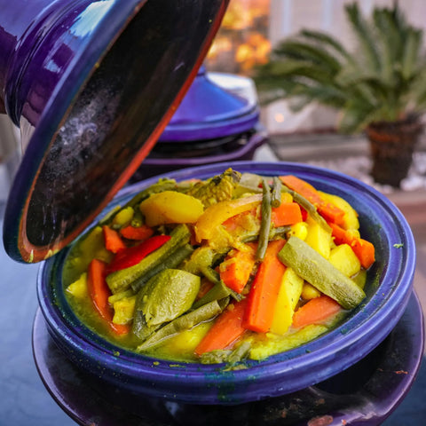 A blue tagine dish brims with vibrant carrots, zucchini, and green beans seasoned with Gustus Vitae’s Fiery Flavors Gourmet Seasoning and Rub Collection. Steam rises as the lid lifts, with a blurred potted plant in the background.