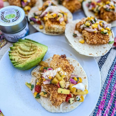 Three soft tacos with breaded fish, corn salsa, and sauce sit with avocado slices. In the background, more tacos and Gustus Vitaes Everything but the Taco Night Collection | Set of 4 rest on a colorful woven placemat.