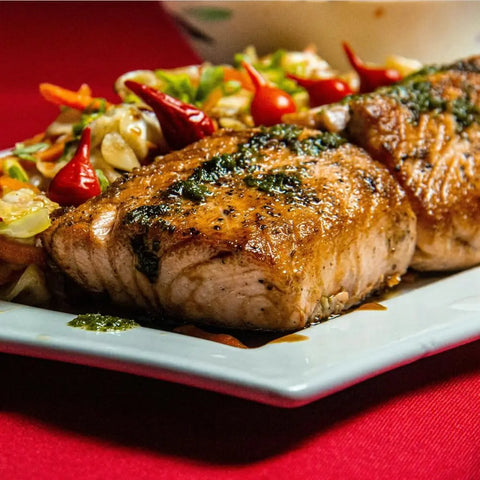 Two grilled salmon fillets with herb sauce sit beside a vibrant veggie salad and red chilies, all seasoned with Gustus Vitaes Grill Master Collection - 4 Pack BBQ seasonings, handmade in the USA, presented on a white plate over a red tablecloth.