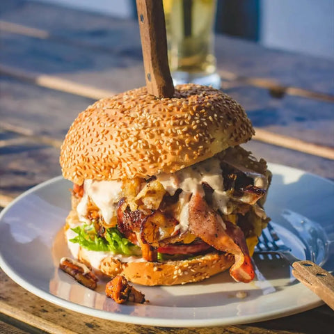 The Grill Master Collection - 4 Pack by Gustus Vitae features gourmet BBQ-seasoned grilled meat, bacon, lettuce, onions, and creamy sauce on a sesame bun. Served on a white plate with a knife through the top bun atop a rustic wooden table.