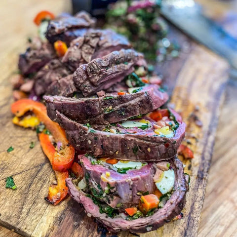 The Grill Master Collection - 4 Pack by Gustus Vitae features juicy steak roulade sliced on a wooden board, stuffed with red peppers, herbs, and veggies. Each tender piece is seasoned with handcrafted spice blends made in the USA. Knife and greens shown in background.