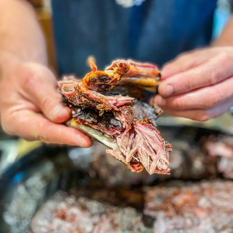 Close-up of a person holding two juicy barbecue ribs, seasoned with Gustus Vitaes Grill Master Collection - 4 Pack, with more meat blurred on a tray in the background.