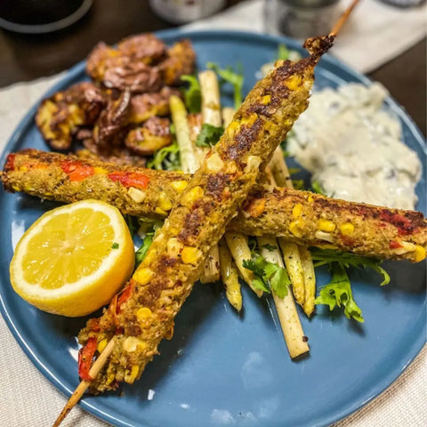 A blue plate features two veggie skewers seasoned with Gustus Vitae’s Complete Kabob Collection, served with white asparagus, leafy greens, fried plantains, creamy sauce, and a lemon half.
