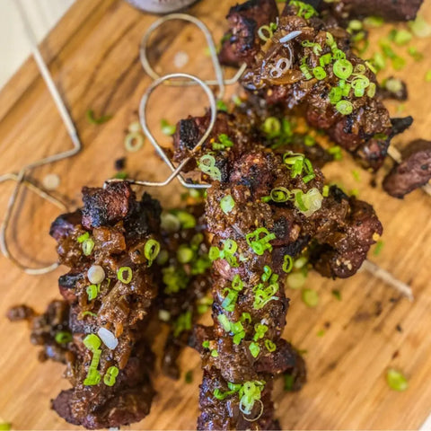 Chargrilled skewered meat, topped with green onions and a rich sauce, served on a wooden board. Rustic twine accents each skewer and highlights the vibrant Middle Eastern flavors of the Complete Kabob Collection by Gustus Vitae.