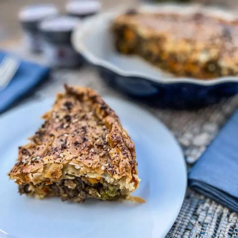 A close-up of a savory pie slice with a golden, seeded crust on a white plate highlights its vegetable and mushroom filling, seasoned to perfection with the Meal Prep Flavor Combo Set - 4 Pack by Gustus Vitae. The rest of the pie sits in the background.