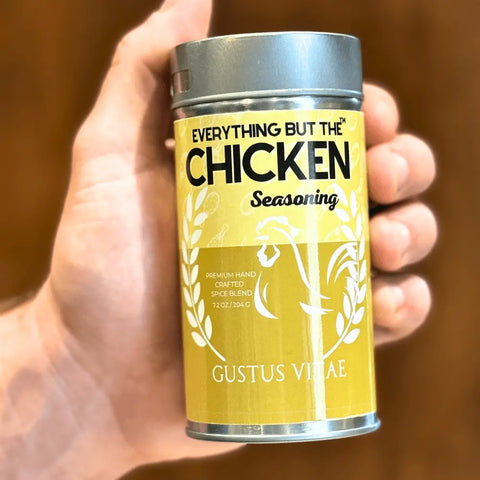 A hand holds a yellow tin from the Gustus Vitae Meal Prep Flavor Combo Set - 4 Pack, featuring a rooster graphic, white text, and a metal lid against a brown blurred background.