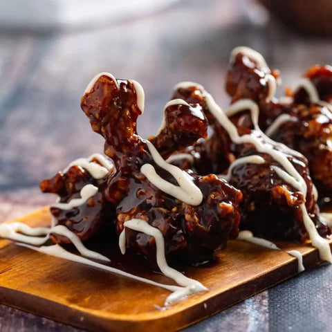 Glazed chicken drumettes coated in Gustus Vitaes Red Hot Wings Seasoning Set and drizzled with creamy white sauce, served on a small wooden board.