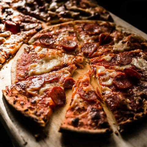 A close-up of a pepperoni pizza cut into slices on wood, topped with melted cheese, crispy edges, and finished with Gustus Vitaes Six Elements of Flavor Seasoning Set, all partially lit by warm lighting.