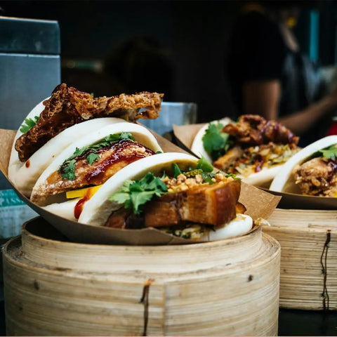 Three steamed bao buns filled with crispy pork belly, fried chicken, cilantro, and vegetables are seasoned with Gustus Vitaes Six Elements of Flavor Seasoning Set and served in bamboo steamers on a market or restaurant counter.
