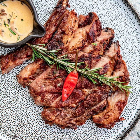 Grilled steak slices on a speckled plate, seasoned with Gustus Vitae’s Gourmet BBQ Steak & Beef Seasoning Gift Set – Six Pack, garnished with rosemary and red chili pepper, served with a side of creamy sauce.
