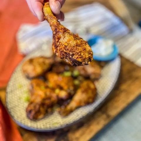 A hand holds a crispy chicken drumstick over a plate with more drumsticks, herbs, and dip—perfectly seasoned with Gustus Vitae’s The Spicy Collection, a set of 4 all-natural BBQ blends from Kitchen, Cooking and Grilling Essentials.
