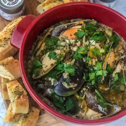 A red pot of mussels in broth, seasoned with Gustus Vitae’s Spicy One Pot Wonders 4 Pack Collection, sits beside herb-topped bread on a wooden tray. A small jar from the gourmet spice set appears in the background.