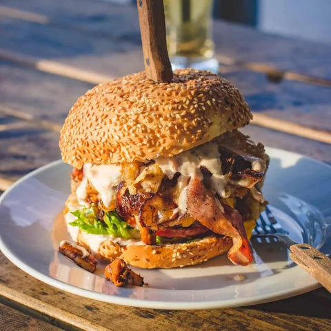 A juicy burger with lettuce, tomato, onions, bacon, and chicken—enhanced using Gustus Vitaes Steak Lovers Gourmet Seasonings & Rubs Collection—served on a white plate outdoors, topped with a knife through the bun.