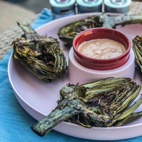Grilled artichoke halves on a pink plate with dipping sauce, served atop a blue cloth and finished with a sprinkle of Gustus Vitae Greatest Hits–Essential Summer Sea Salts | Set of 4.