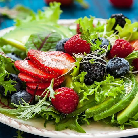 A salad of mixed greens, berries, and avocado is arranged on a white plate, finished with Gustus Vitae’s Greatest Hits- Essential Summer Sea Salts set for vibrant color and fresh flavor.