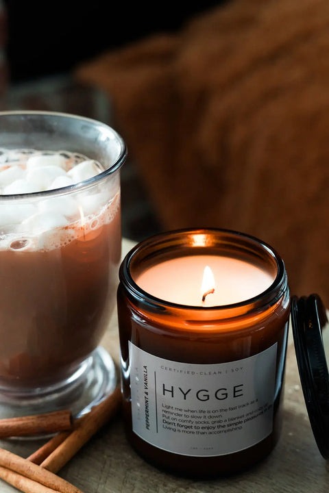 A glass mug of hot chocolate with marshmallows sits next to a lit Shanti + Tula HYGGE candle (Peppermint + Vanilla, 100% soy wax) on a wooden surface, with cinnamon sticks nearby for a cozy, eco-friendly vibe.