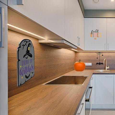 A modern kitchen is decorated for Halloween with a pumpkin, bats, and the Chatelet Manufacturing Halloween Drink Up Witches Wall Sign (9.5 x 6.5) above white cabinets for a festive touch.