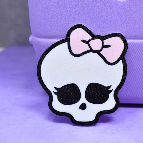 The FRESHe BAGLETS Halloween Cute Pink Bow & Skull Charm by Chatelet Manufacturing features a cartoon white skull with big black eye sockets, long lashes, and a pink bow—perfect for accessorizing any rubber tote bag. Made in USA.