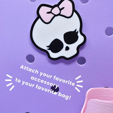 A white skull with long eyelashes and a pink bow is attached to a lavender pegboard. Text: “Attach your favorite FRESHe BAGLETS - Halloween Cute Pink Bow & Skull Charm by Chatelet Manufacturing to your rubber tote bag!”.