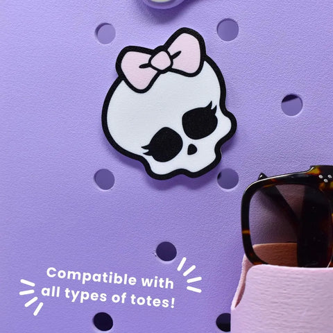 The FRESHe BAGLETS Halloween Cute Pink Bow & Skull Charm by Chatelet Manufacturing is shown on a lavender pegboard. Compatible with all rubber tote bags, its the perfect cute Halloween accessory. Pink holder with tortoiseshell sunglasses nearby.