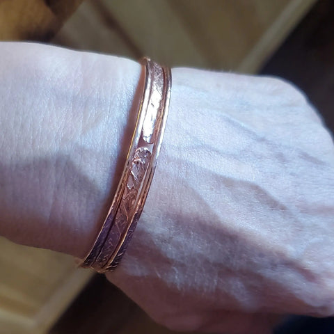 A close-up shows a wrist wearing two thin Stackable Hammered Double Canted Angle Bangles by Alexa Martha Designs against a light background.