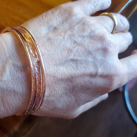 A close-up shows a hand wearing Alexa Martha Designs’ Stackable Hammered Double Canted Angle Bangles with a gold ring; the textured copper bangles stand out against wood and glass, highlighting natural skin details.