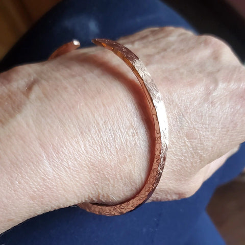 A close-up shows a hand wearing an Alexa Martha Designs Stackable Hammered Double Canted Angle Bangle, featuring a textured, open-ended handmade look with a hammered finish. The person’s skin and dark blue fabric appear in the background.