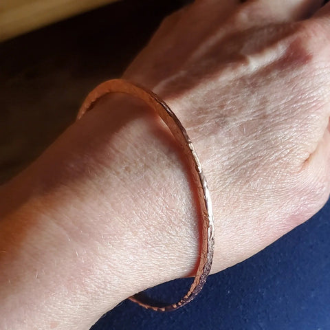 A close-up of a wrist wearing an Alexa Martha Designs Stackable Hammered Double Canted Angle Bangle, a thin handmade copper bracelet perfect for stacking with other bangles, set against a blurred background.